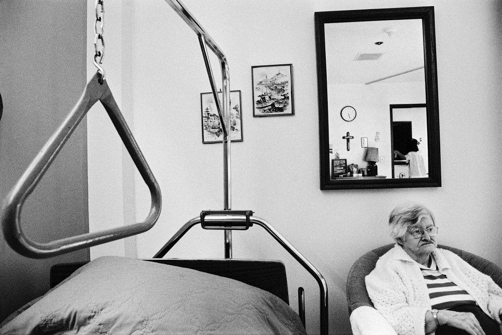 An elderly person sitting in a chair beside a hospital bed, with a mirror reflecting the room
