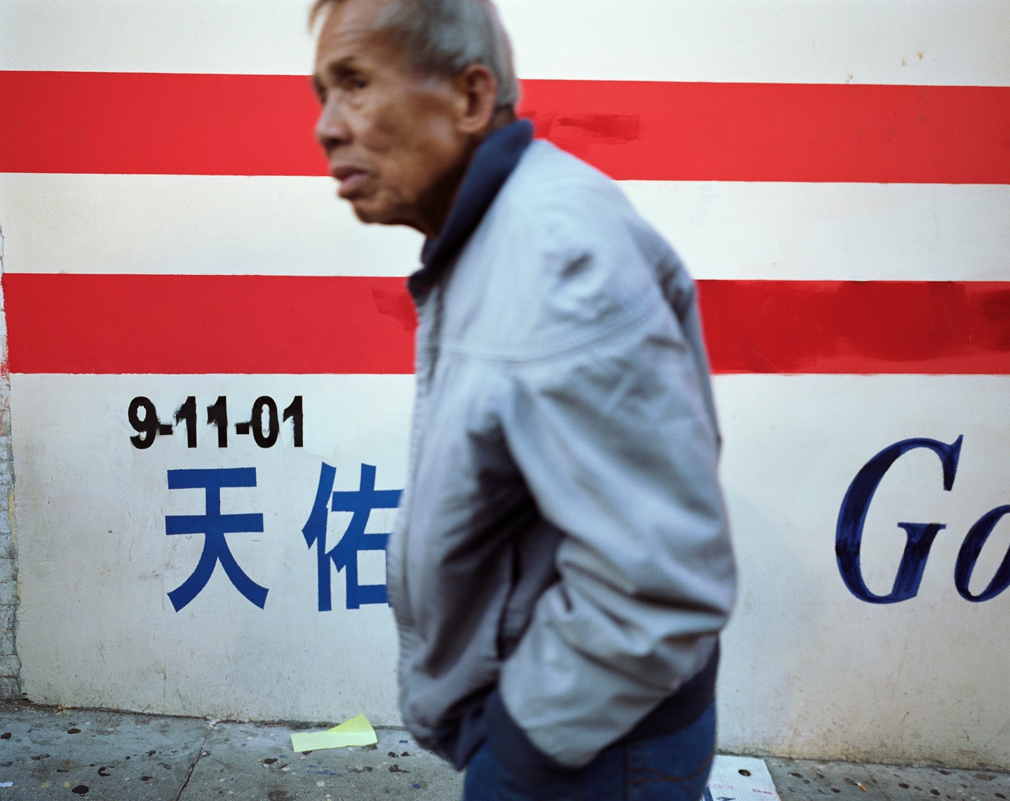 An elderly man walking past a wall with red stripes and writings in English and Chinese