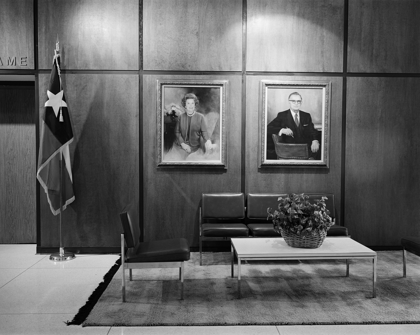 Interior of a room featuring a flag, two portraits on the wall, minimalist furniture, and a potted plant