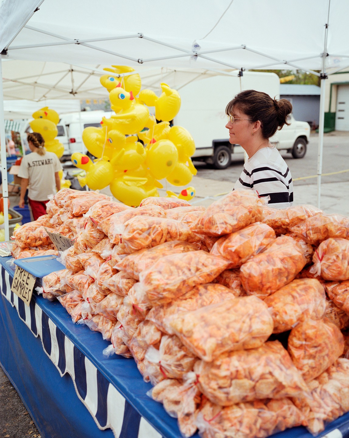 A vendor at a market stands next to bags of snacks and a display of yellow rubber duck balloons