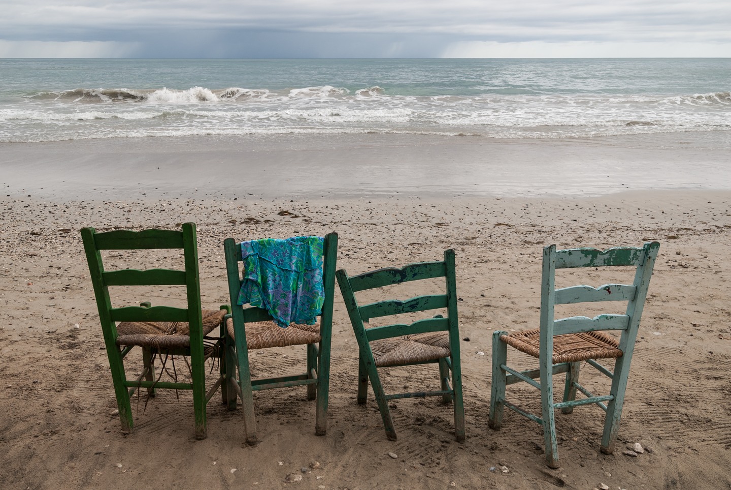 Four weathered chairs in varying colors facing the ocean on a sandy beach