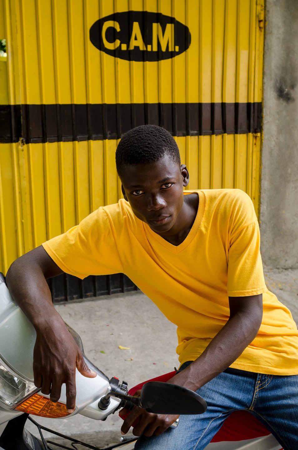 A young man in a yellow shirt leans casually on a scooter in front of a bright yellow wall