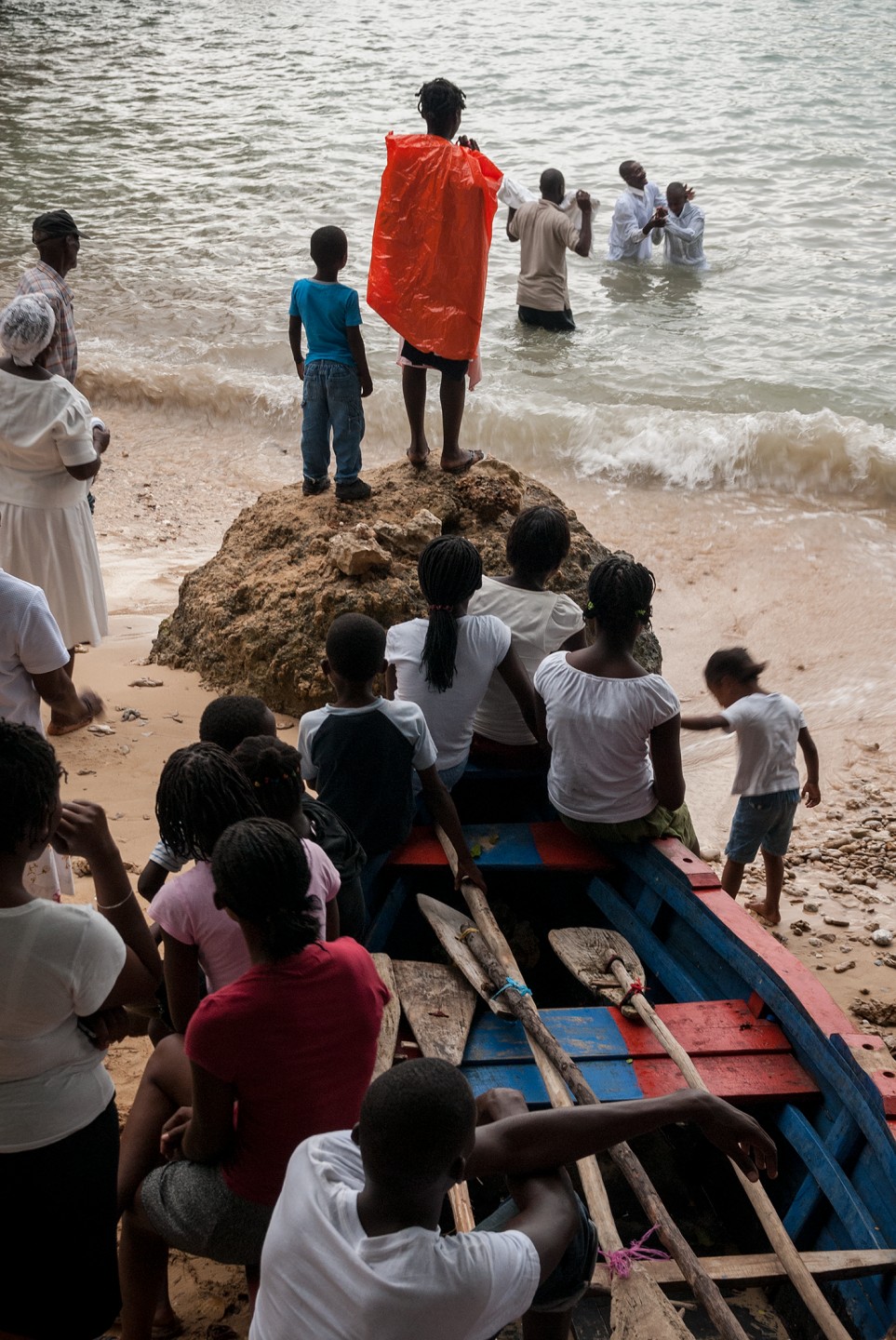 A group of people gathers at the beach, some on a boat and others on rocks, with one child wearing a bright orange cape