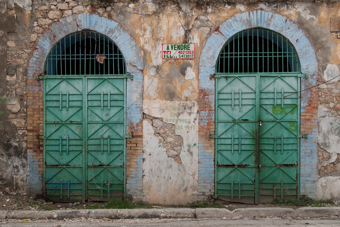 Weathered brick wall with two green arched doors and a "For Sale" sign