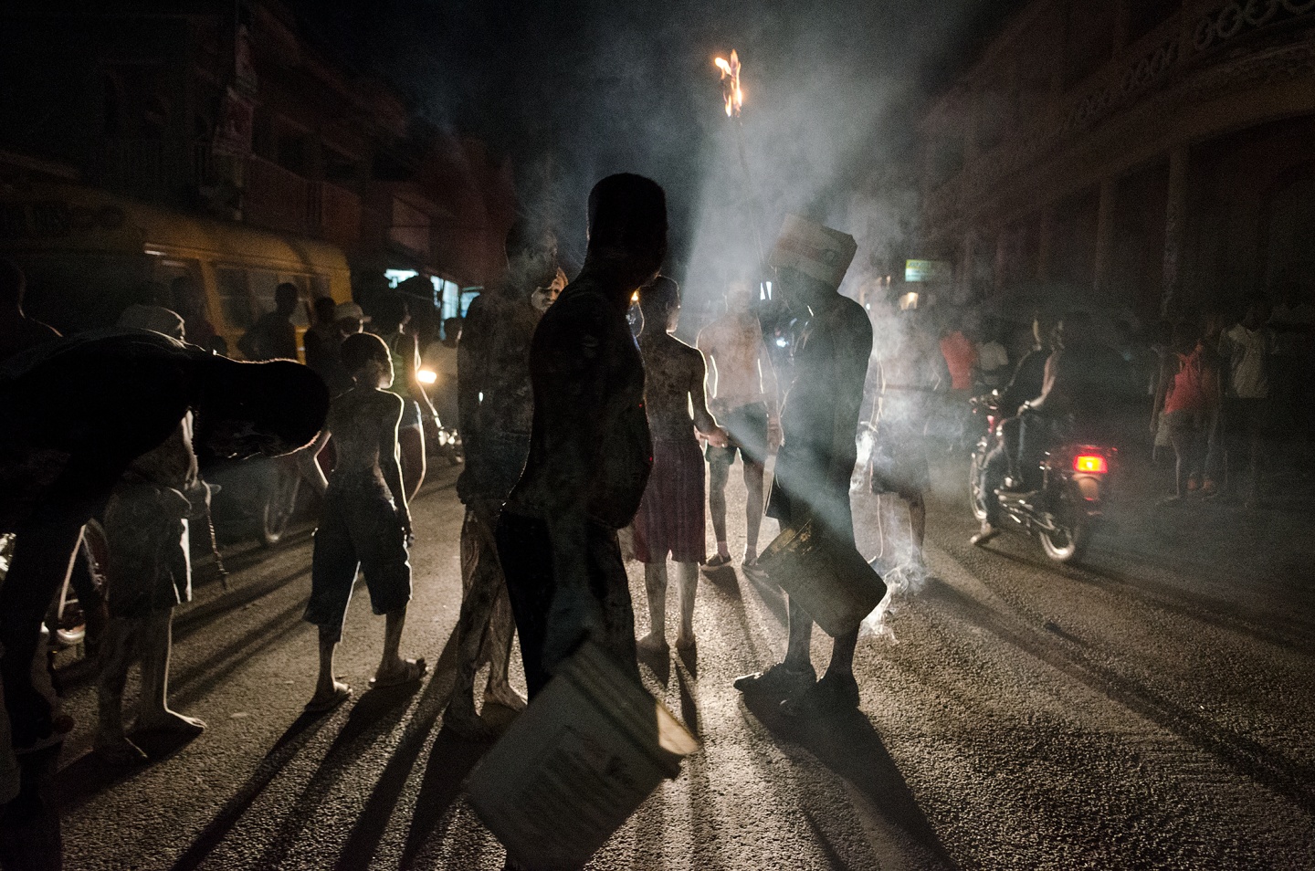 People gathered on a dimly lit street, surrounded by smoke and shadows, during a nighttime celebration
