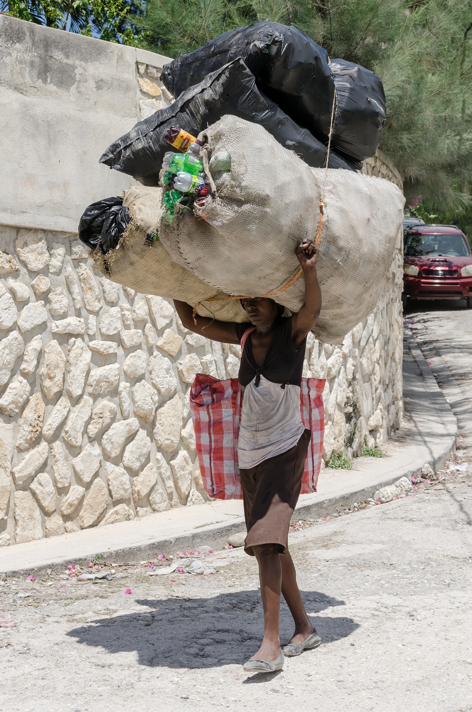 A person carries a large bag of recyclables on their back along a narrow street