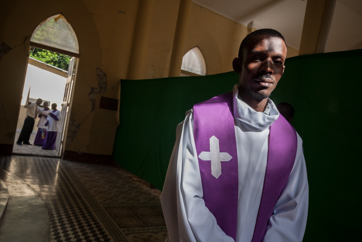 A priest in a purple stole stands in a church with congregants visible in the background