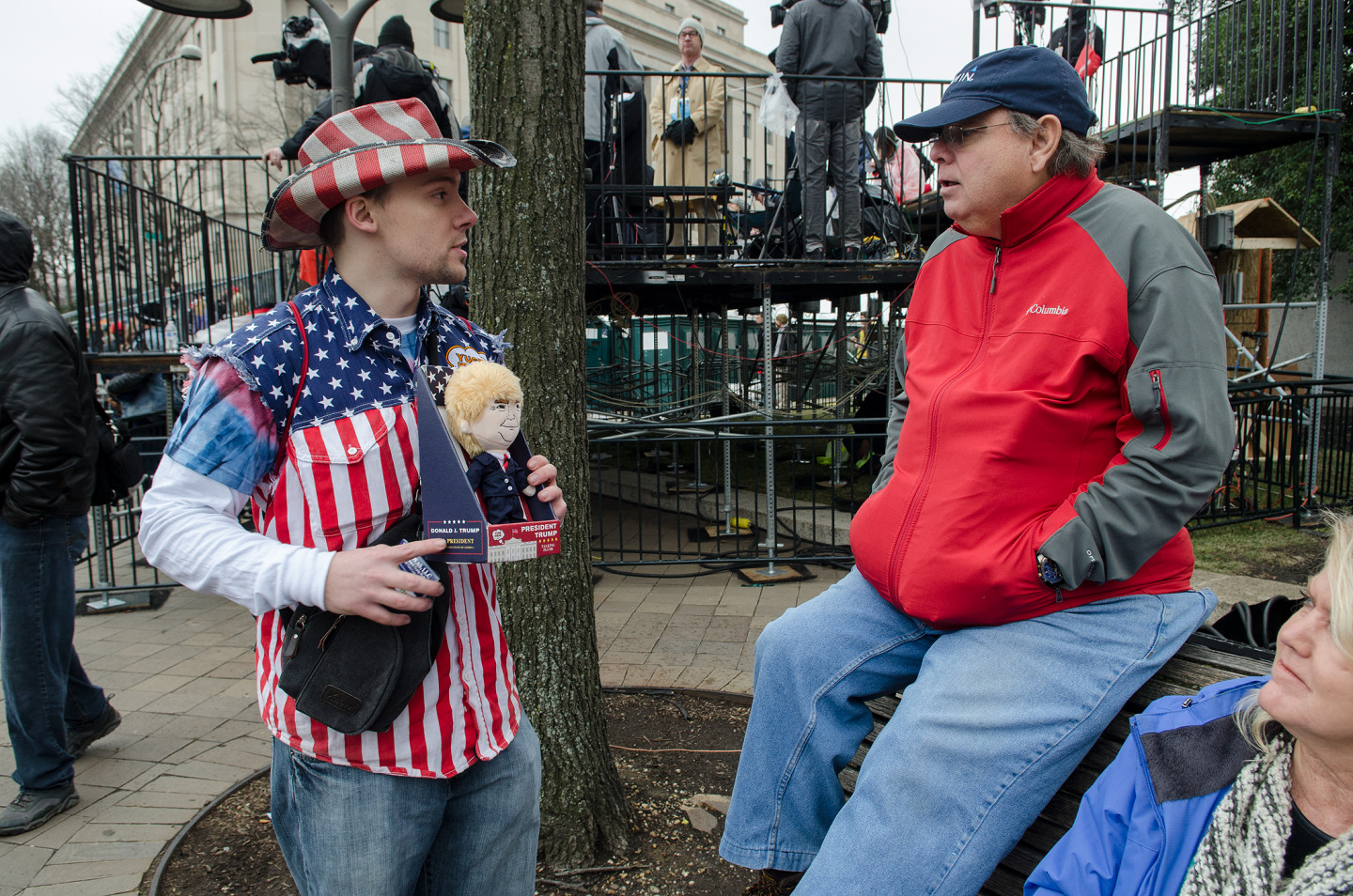 Man in a patriotic outfit holding a doll engages in conversation with another man in a red jacket
