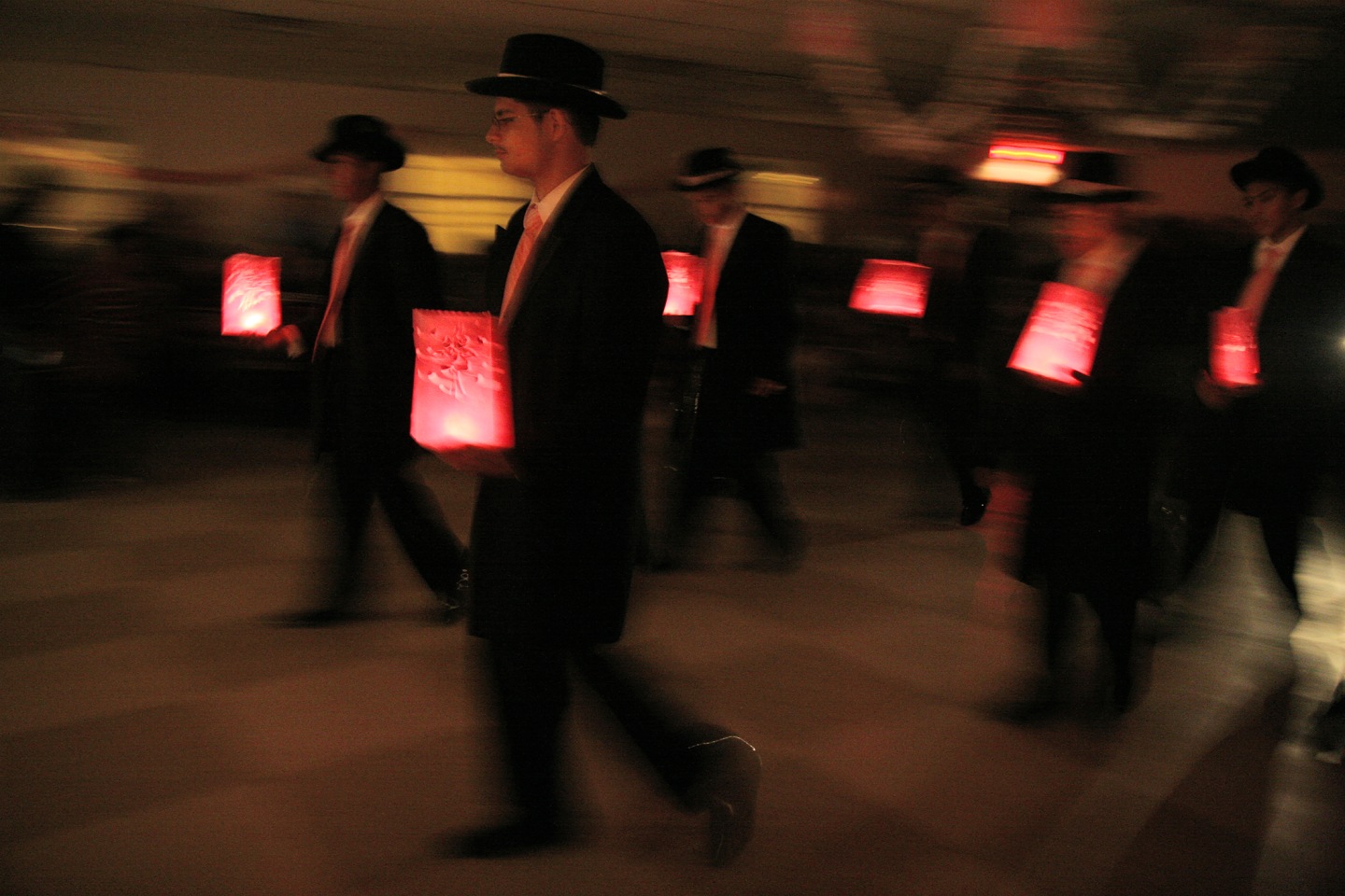Men in dark suits and hats walking while holding glowing red lanterns