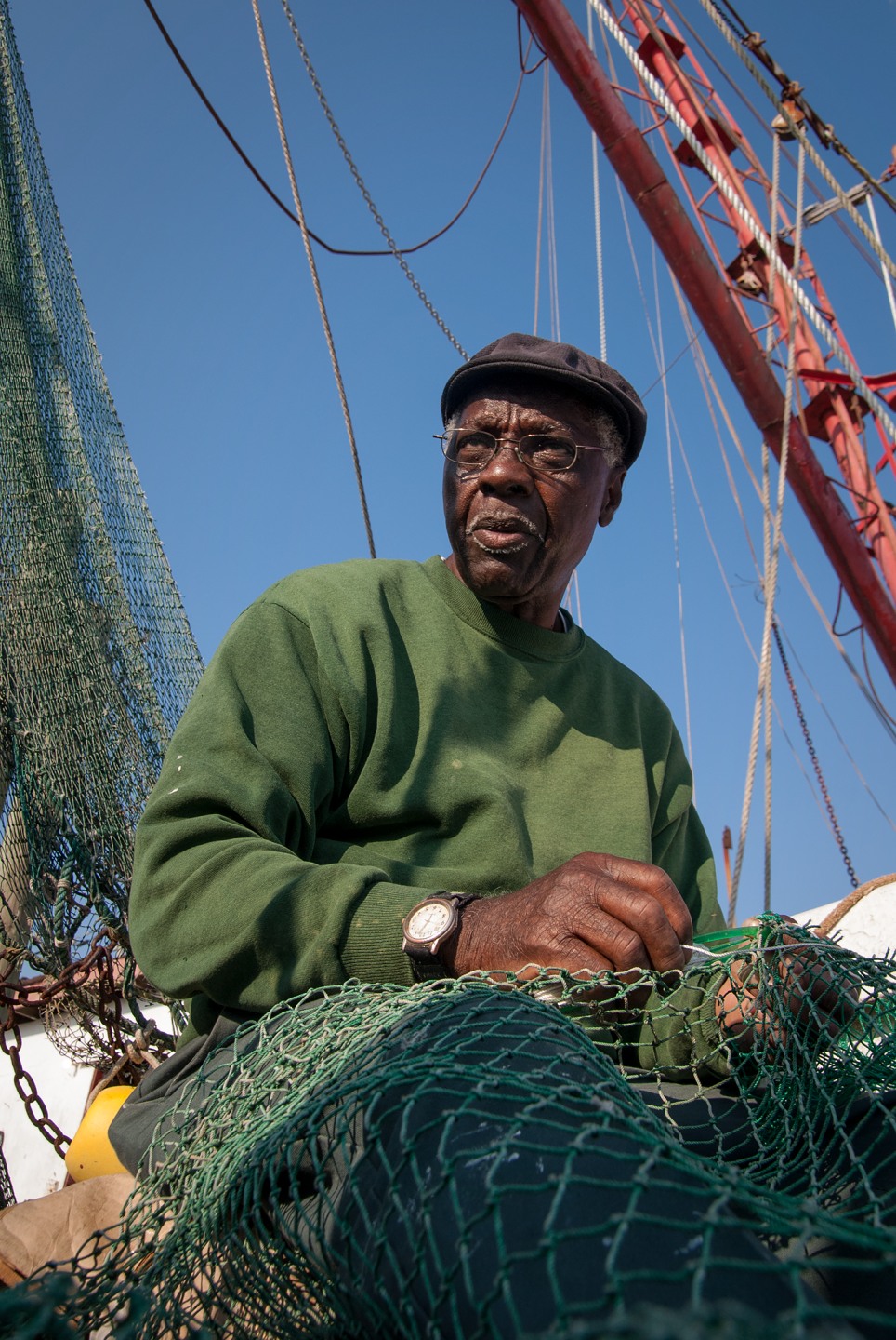 A man in a green sweater and cap sits on a fishing boat, working with netting against a clear blue sky