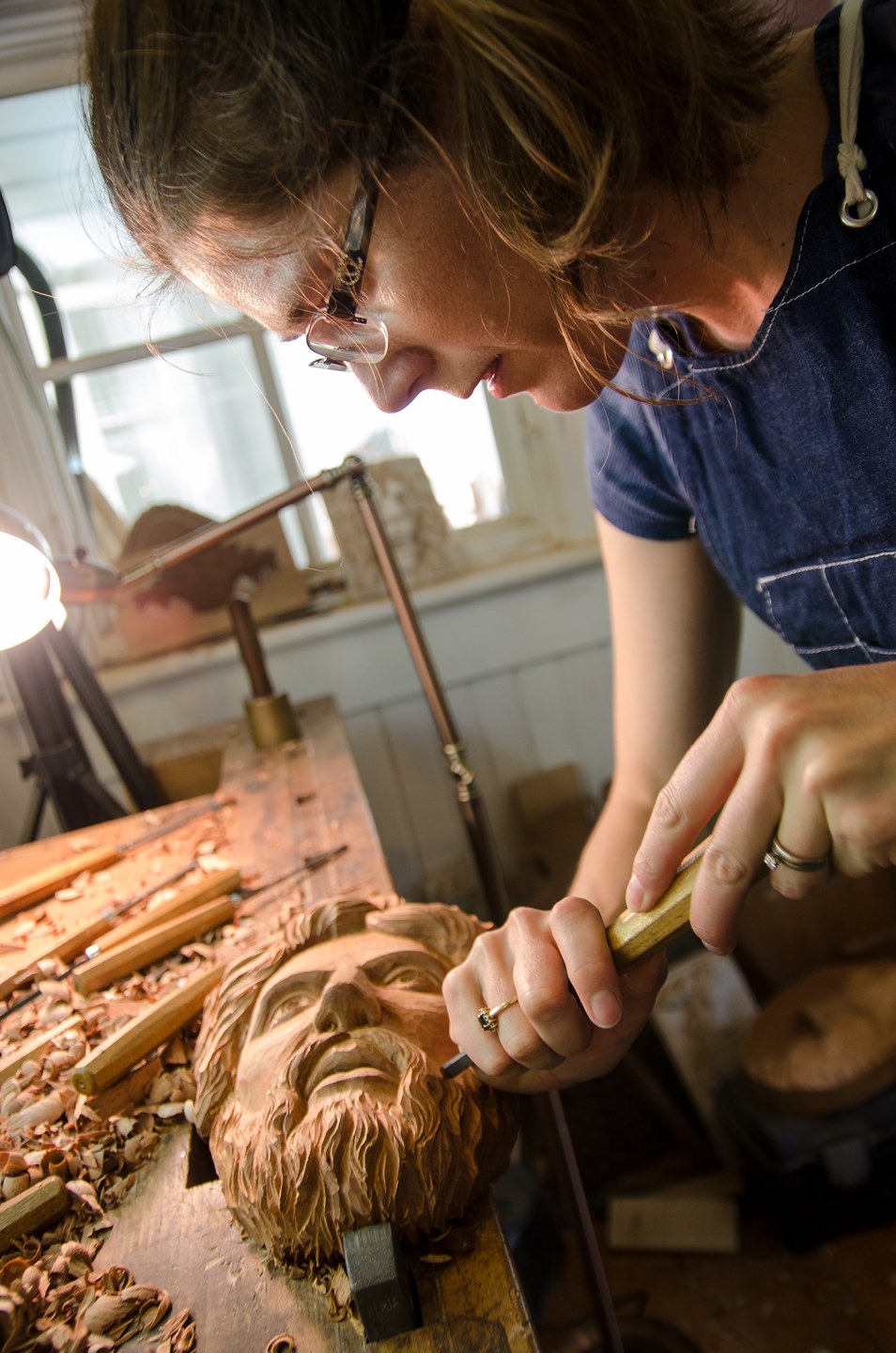 A woman carves a wooden sculpture of a bearded face in her workshop