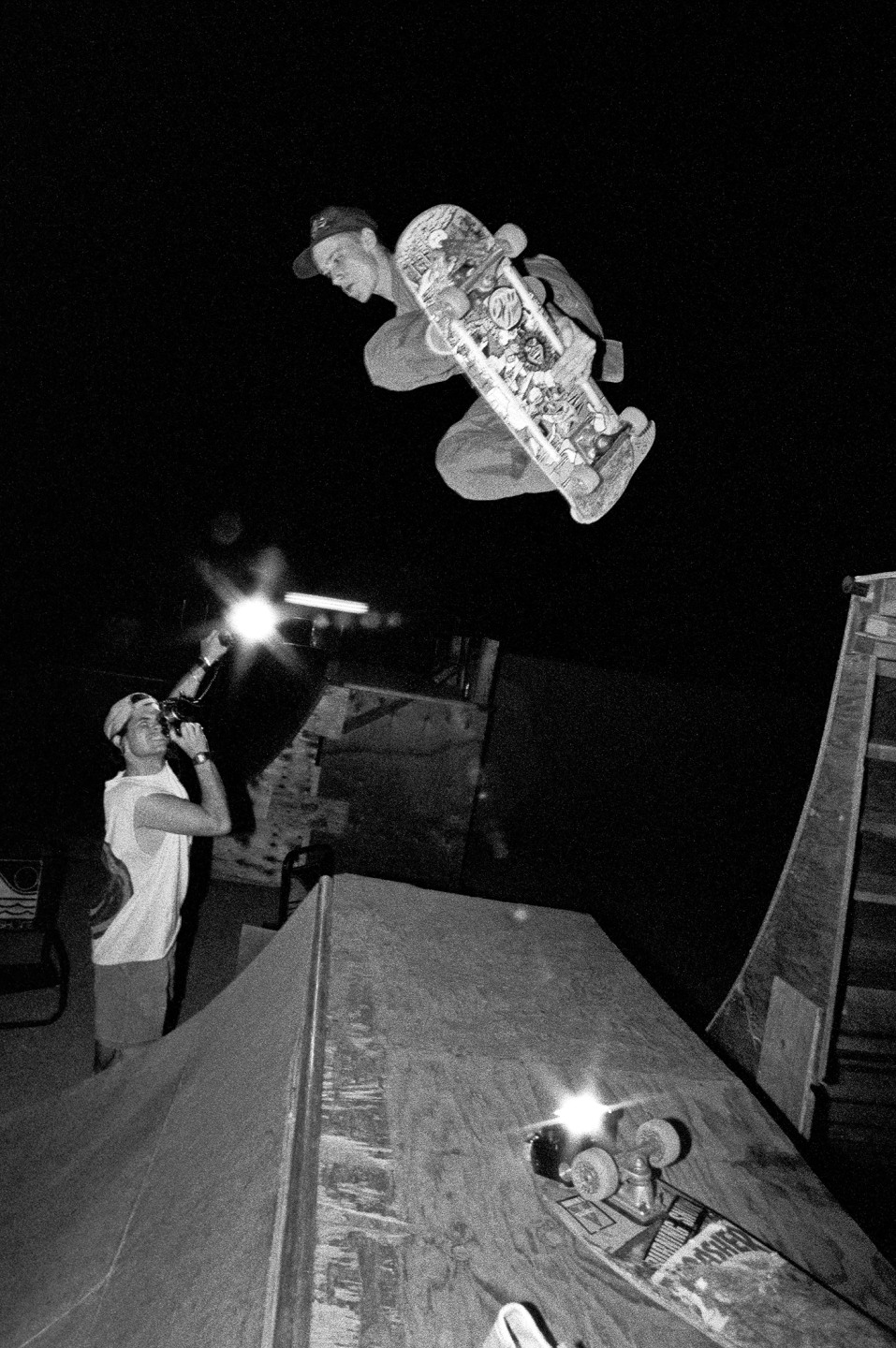 Skateboarder performing a trick in mid-air at night while another person films with a flashlight