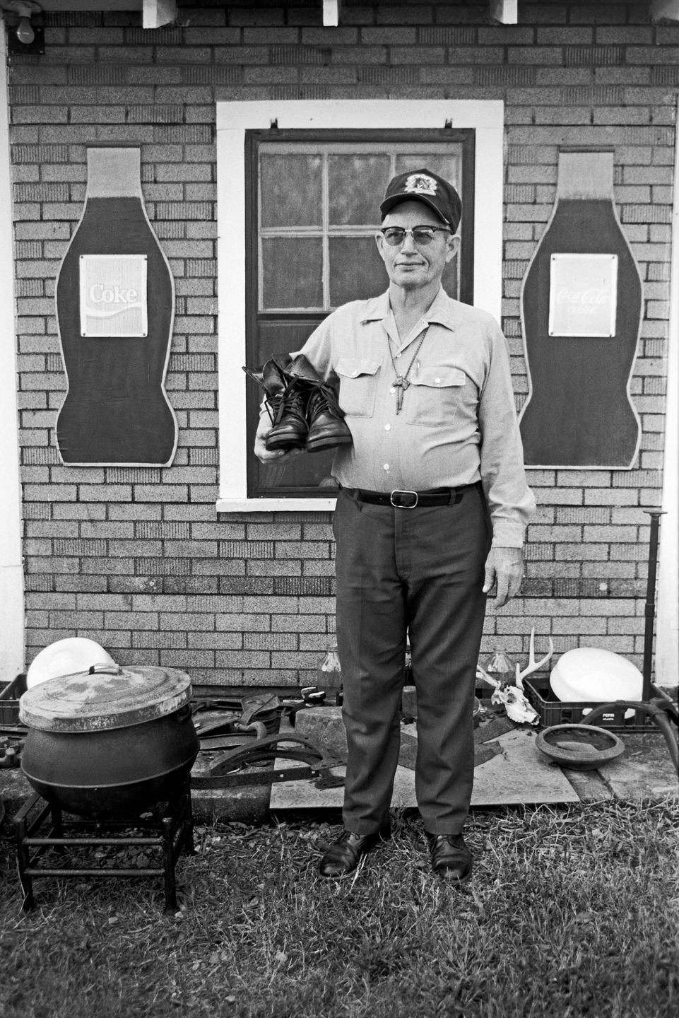 Man in a cap holding a pair of boots in front of a brick wall with Coca-Cola advertisements