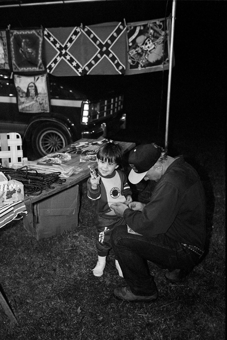 A child sits at a table with various items while an older man adjusts the child's clothing at a nighttime outdoor gathering