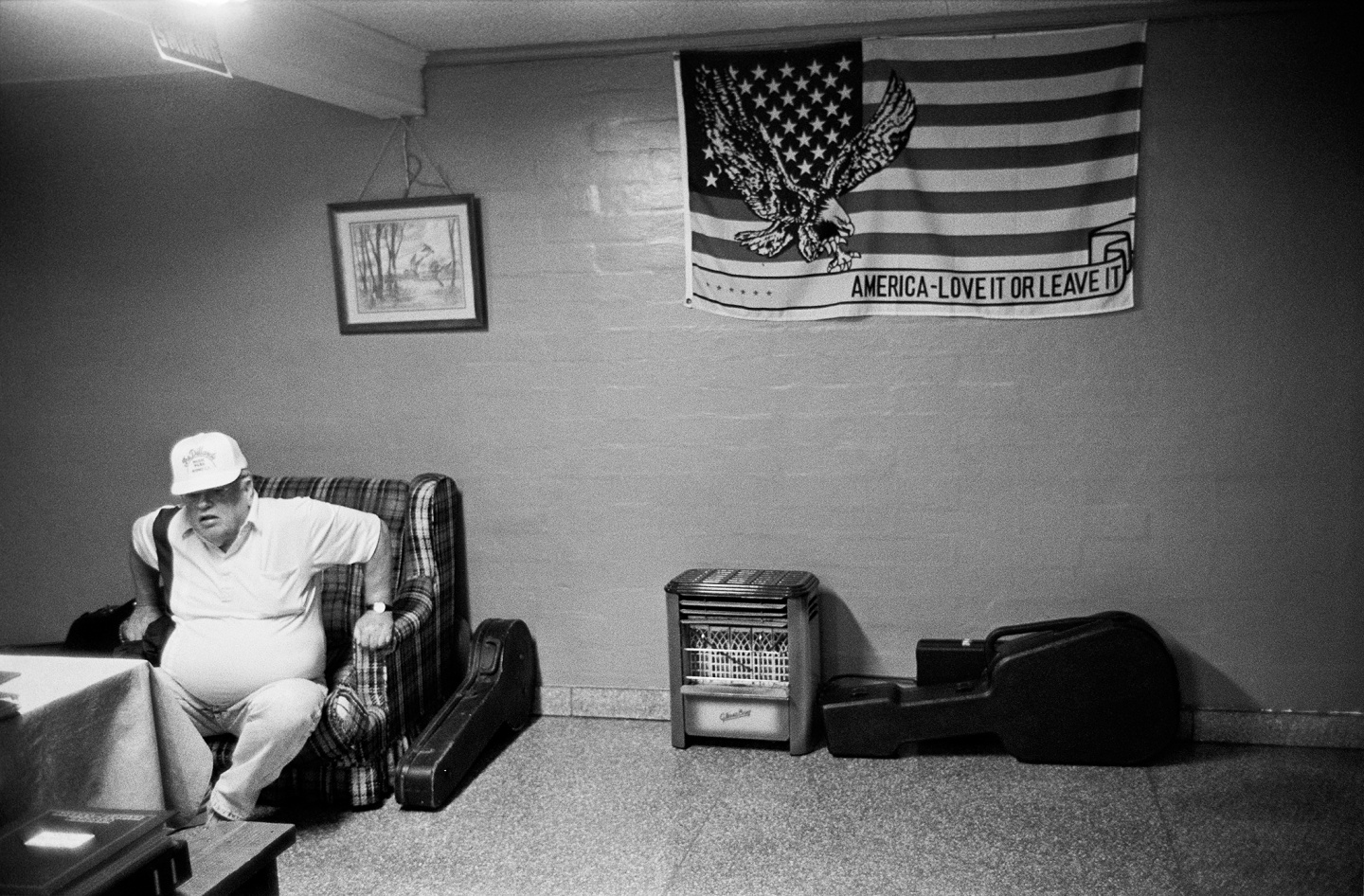 A man sits on a couch in a dimly lit room adorned with an American flag and a painting