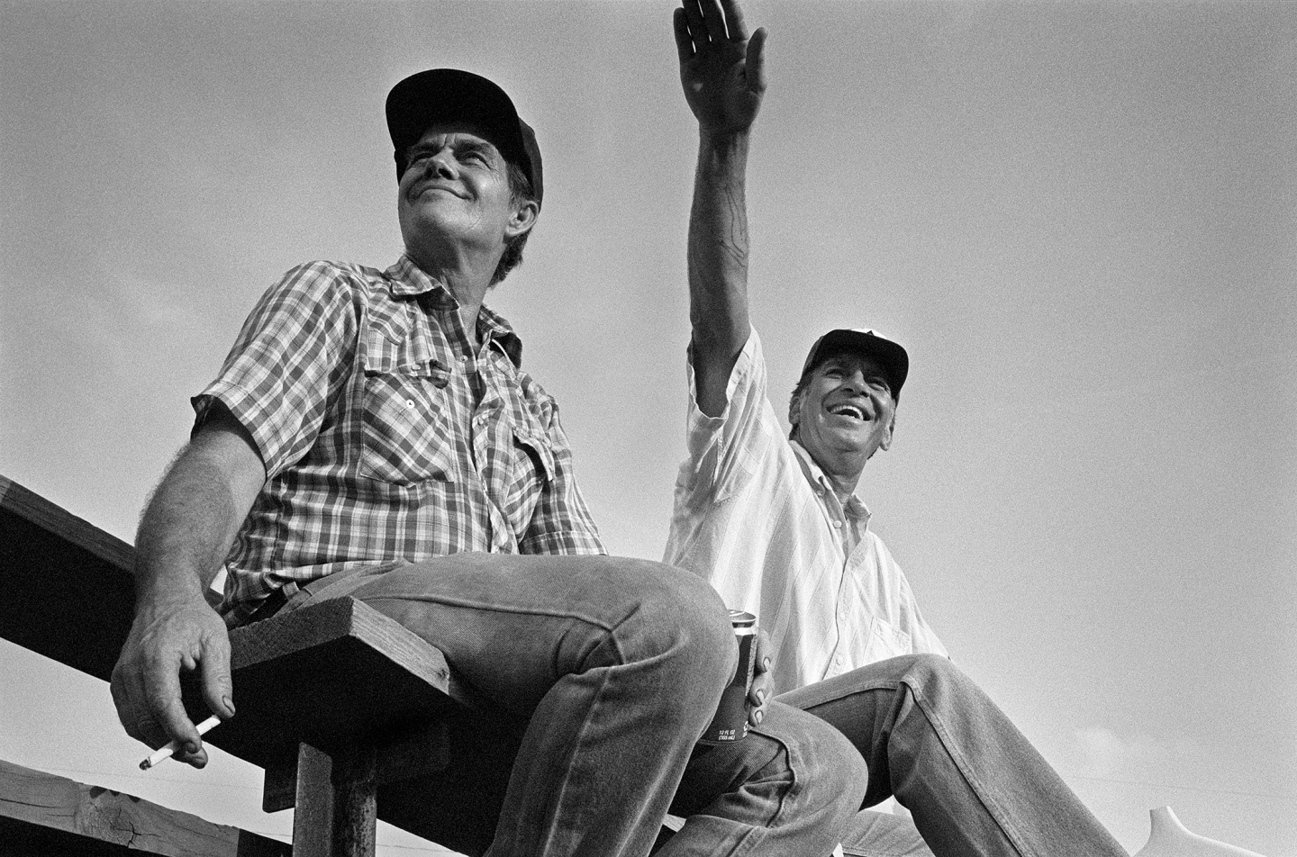 Two men sitting on a wooden bench smiling and gesturing, in a black and white photograph