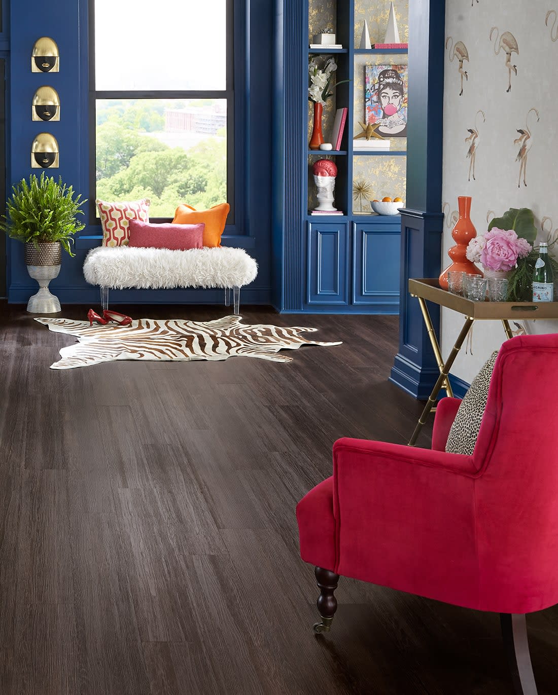 This living room shows new furniture including a soft textured red chair, padded acrylic bench, faux fur rug, and stylish metallic accents. With Lovina Beach waterproof vinyl flooring.