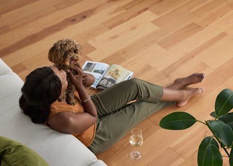 7/16 in x 5.4 in Matte Natural Hickory Engineered Hardwood Flooring closed up of woman sitting on floor leaning against sofa holding dark brown small dog with wine glass and magazine on floor