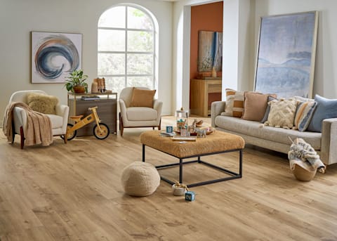 5mm with Pad Yorkshire Oak Waterproof Rigid Vinyl Plank Flooring in living room with beige furnishings plus gold upholstered ottoman and wooden tricycle and knotting material in a basket