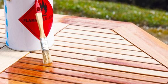 Eine Dose mit Holz-Pflegeöl und ein Pinsel stehen auf einem Teakholztisch.
