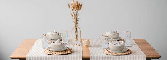 Tavolo in legno chiaro con runner a righe beige, ciotole tono su tono e bicchieri trasparenti. Al centro, un vaso con fiori secchi dona un tocco naturale ed elegante.