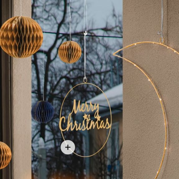 Deko-Hänger in gold mit Inschrift "Merry Christmas", aufgehangen vor einem Fenster. Daneben hängen blaue und braune Weihnachtskugeln von der Decke.