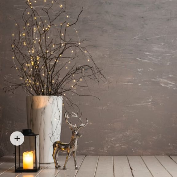 Eine schwarze Laterne mit LED-Kerze steht neben einem silbernen Deko-Hirsch und einer großen marmorierten Vase gefüllt mir Zweigen und einer Lichterkette.