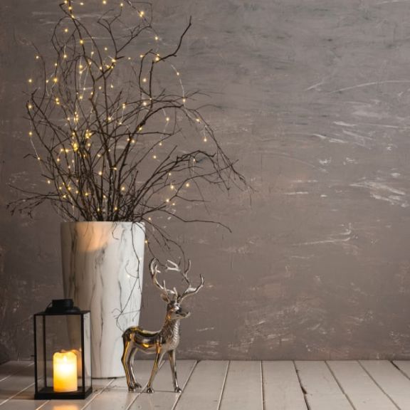Eine schwarze Laterne mit LED-Kerze steht neben einem silbernen Deko-Hirsch und einer großen marmorierten Vase gefüllt mir Zweigen und einer Lichterkette.