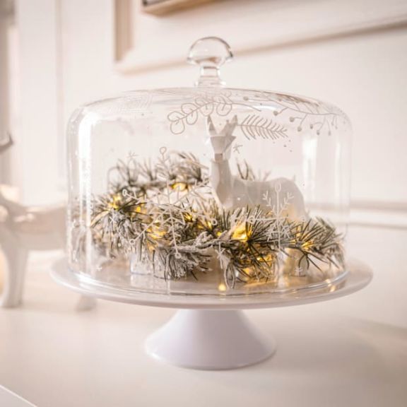 Cloche auf Tortenplatte, innen dekoriert mit einer weißen Rehfigur und einer leuchtenden Zweiggirlande.