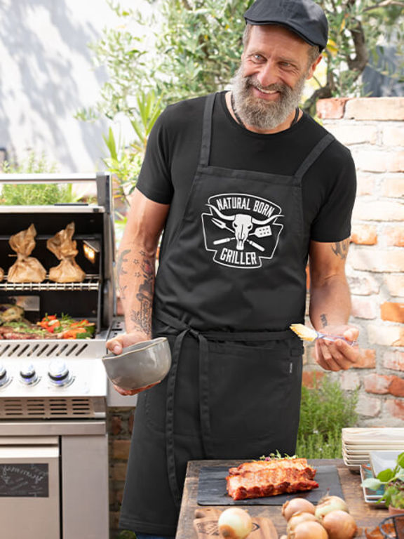 Mann mit "Natural Born Griller" Schürze vor einem Grill