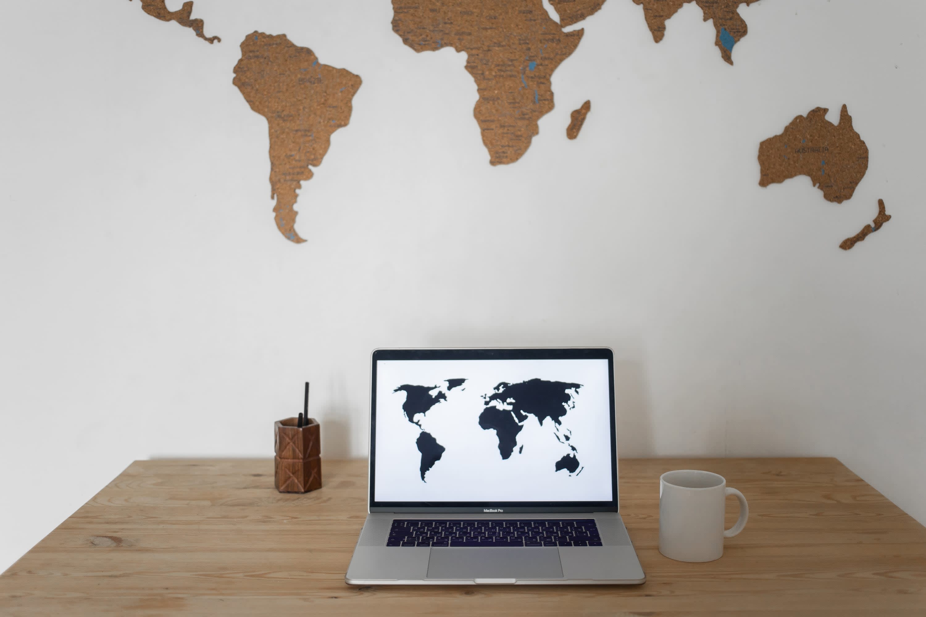 A white cup, pen holder, and laptop with a world map image on a brown table represent real estate's global reach.