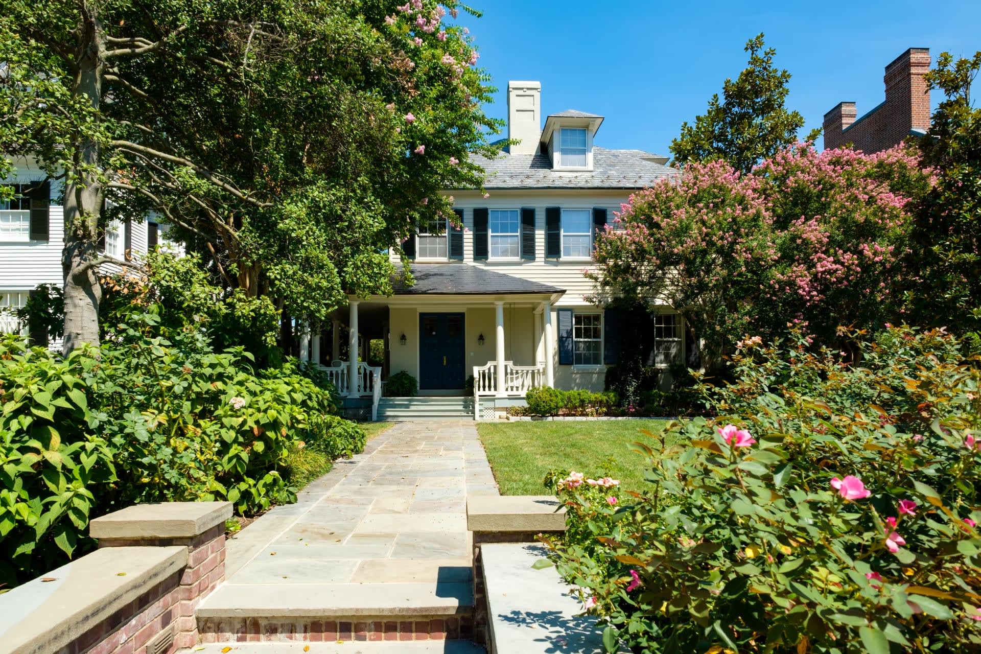 A white Colonial-style home with a front porch, a white picket fence, a brick pathway, and a lush front yard.