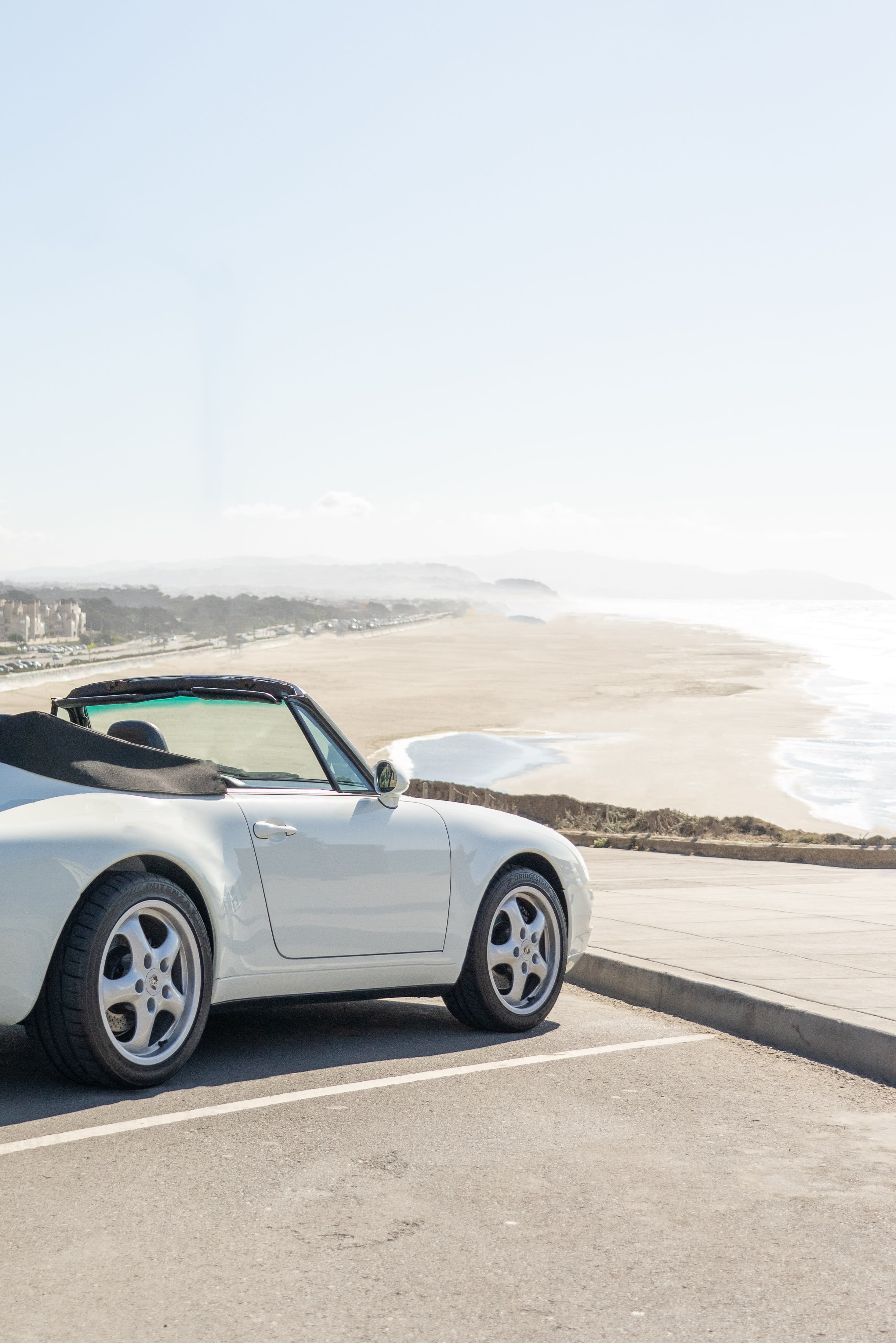 A white convertible car with its top down, parked on a sandy beach next to the ocean.