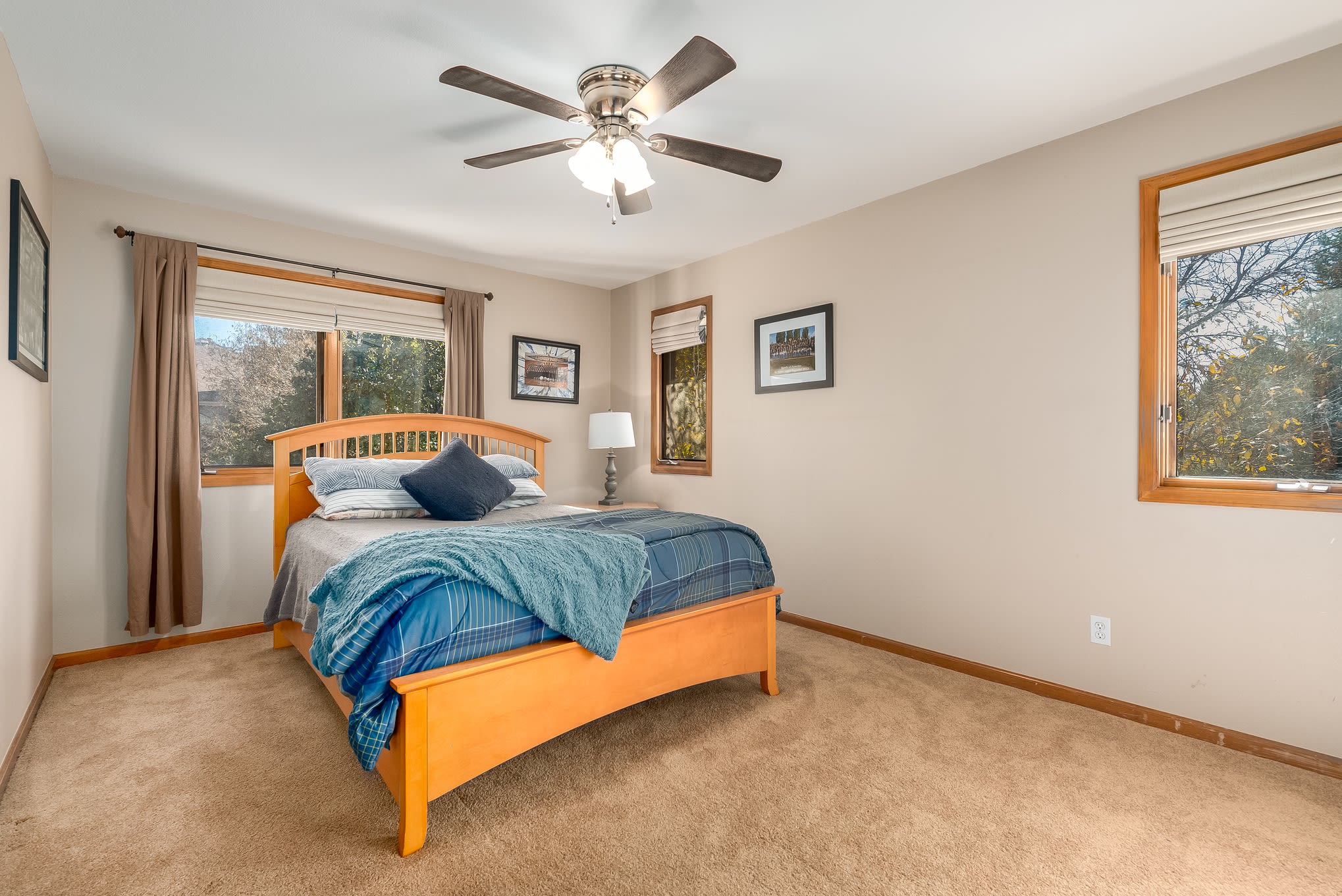 Bedroom with a full-size bed, light comforter, wooden nightstand, white-shade lamp, ceiling fan, and outside-view windows.