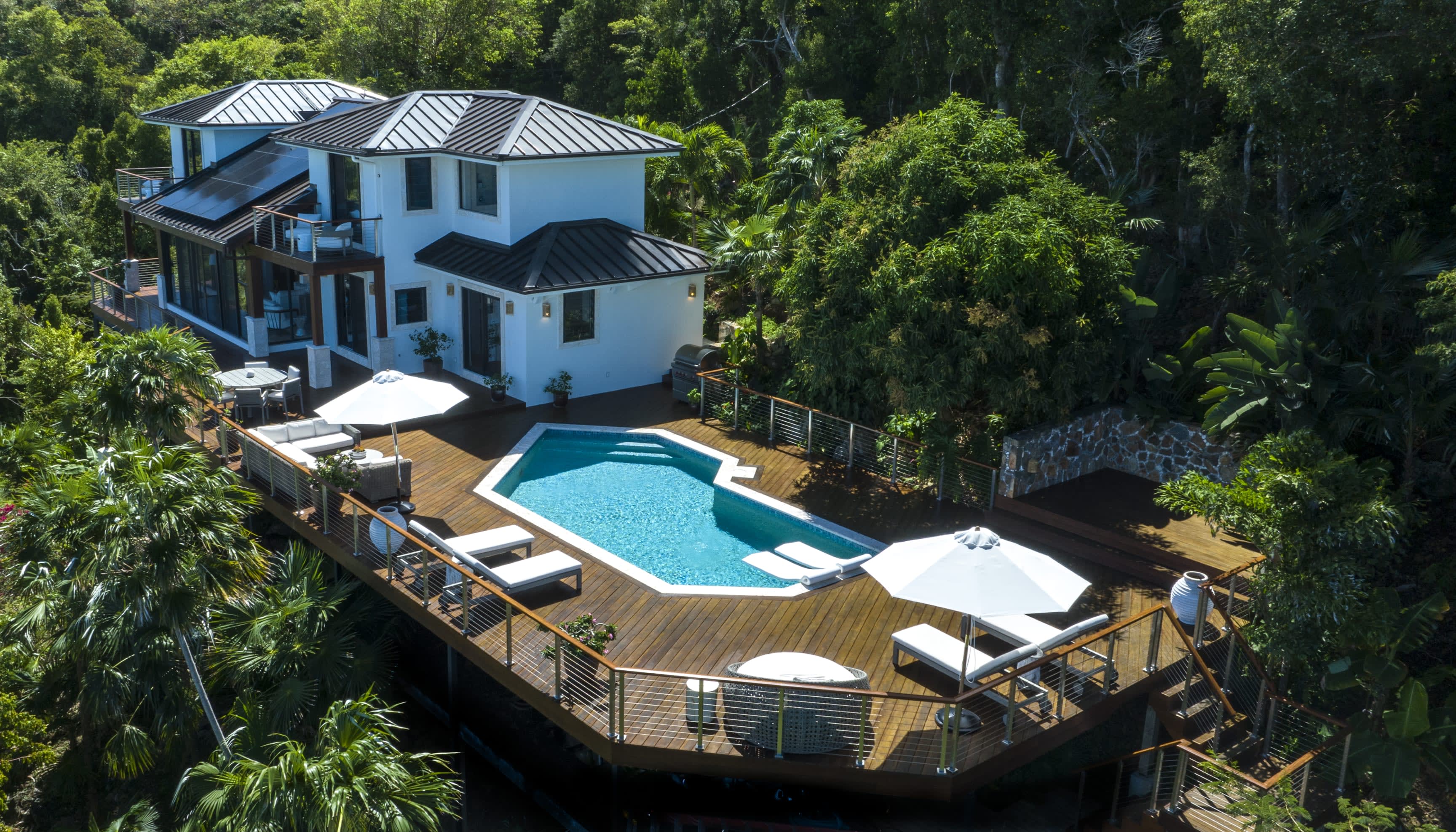 An aerial view of a modern house with a swimming pool on a wooden deck, surrounded by lush greenery