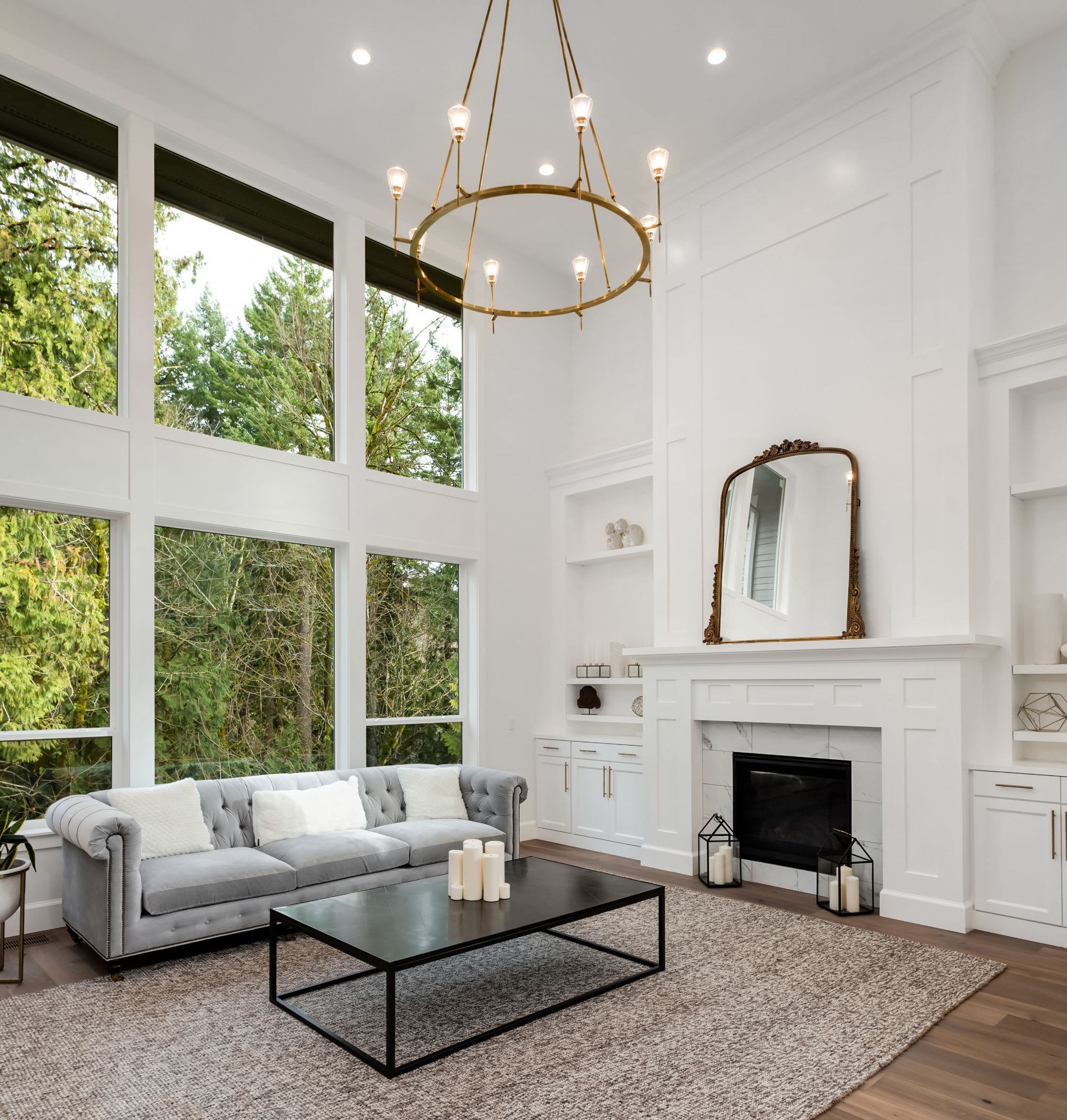  A bright and luxurious living room featuring a grand, tall ceiling and large windows that offer a view of lush green trees outside.