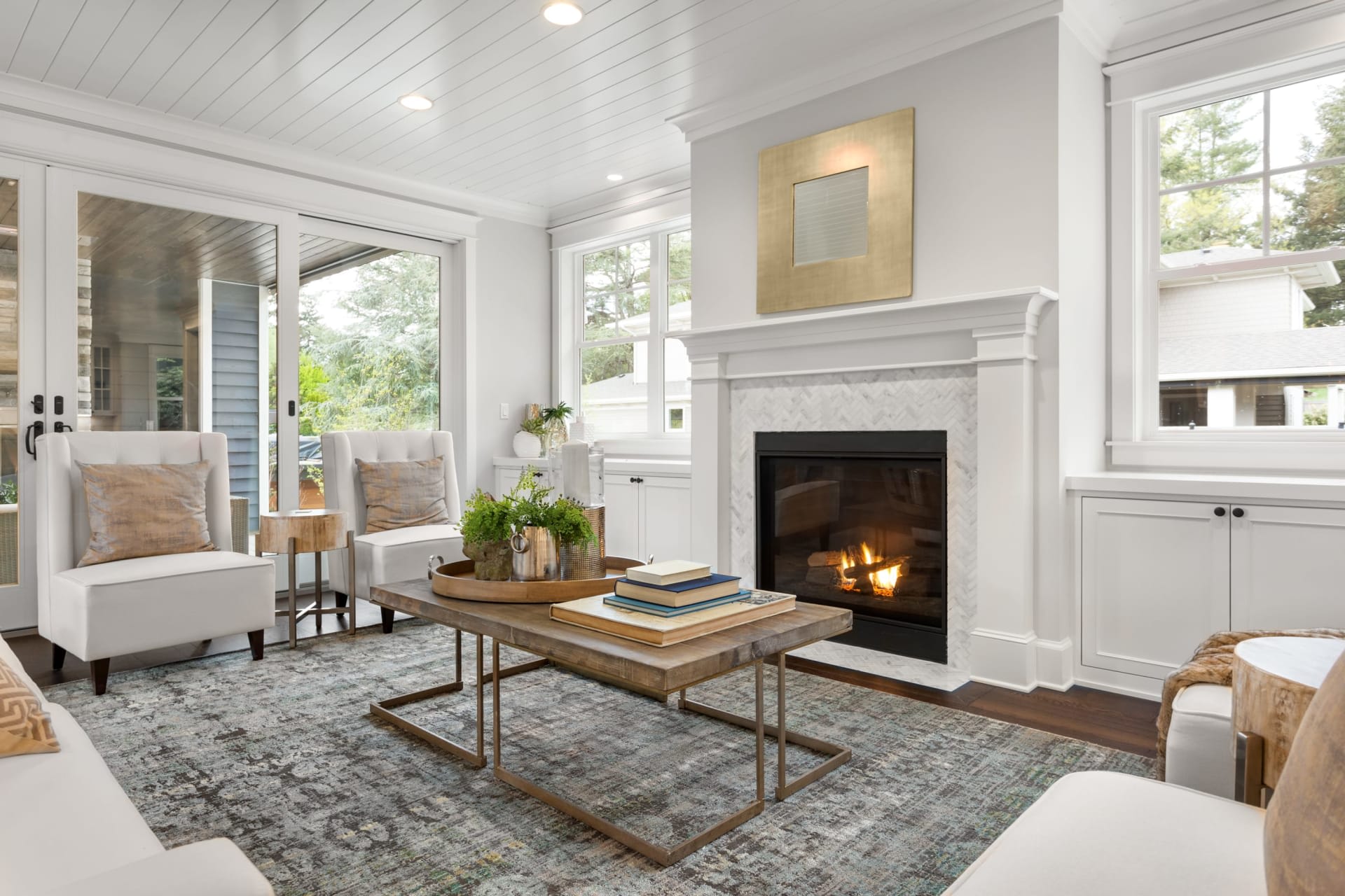 A cozy living room with white furniture, a coffee table, a fireplace, a glass door, and windows with an outdoor view.