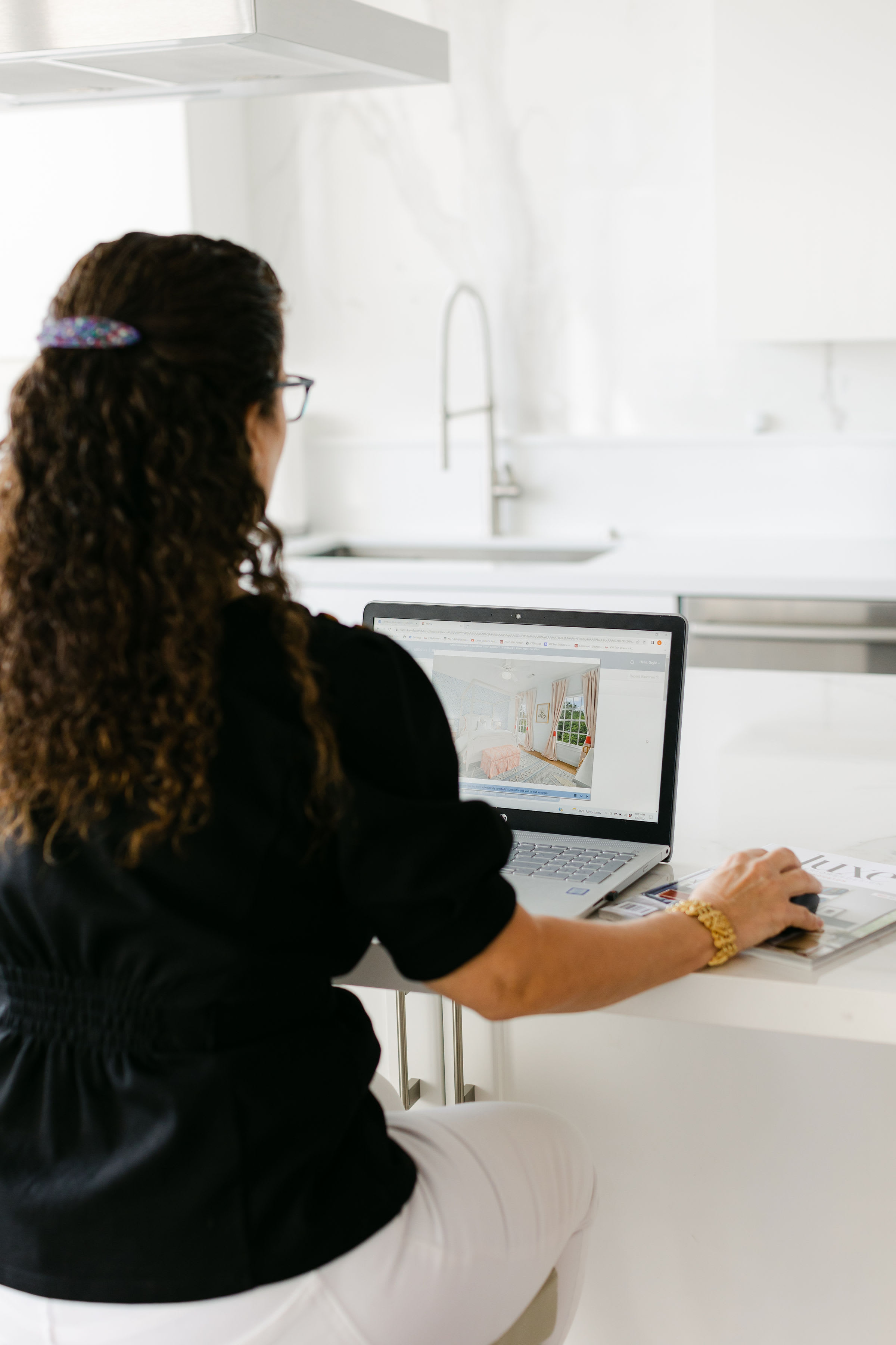 In a bright, modern kitchen, a woman in a black blouse and white pants browses luxury homes on her laptop.