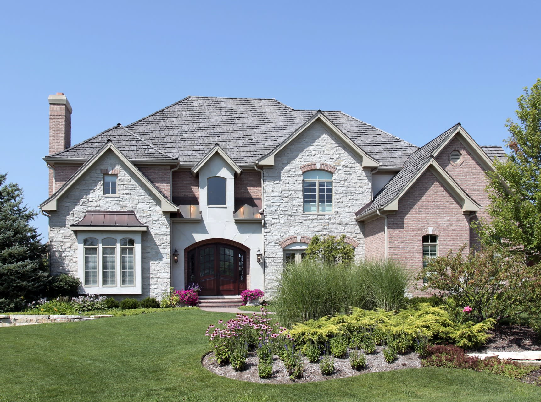 A large brick house with a slate roof and a large front yard filled with flowers and plants.