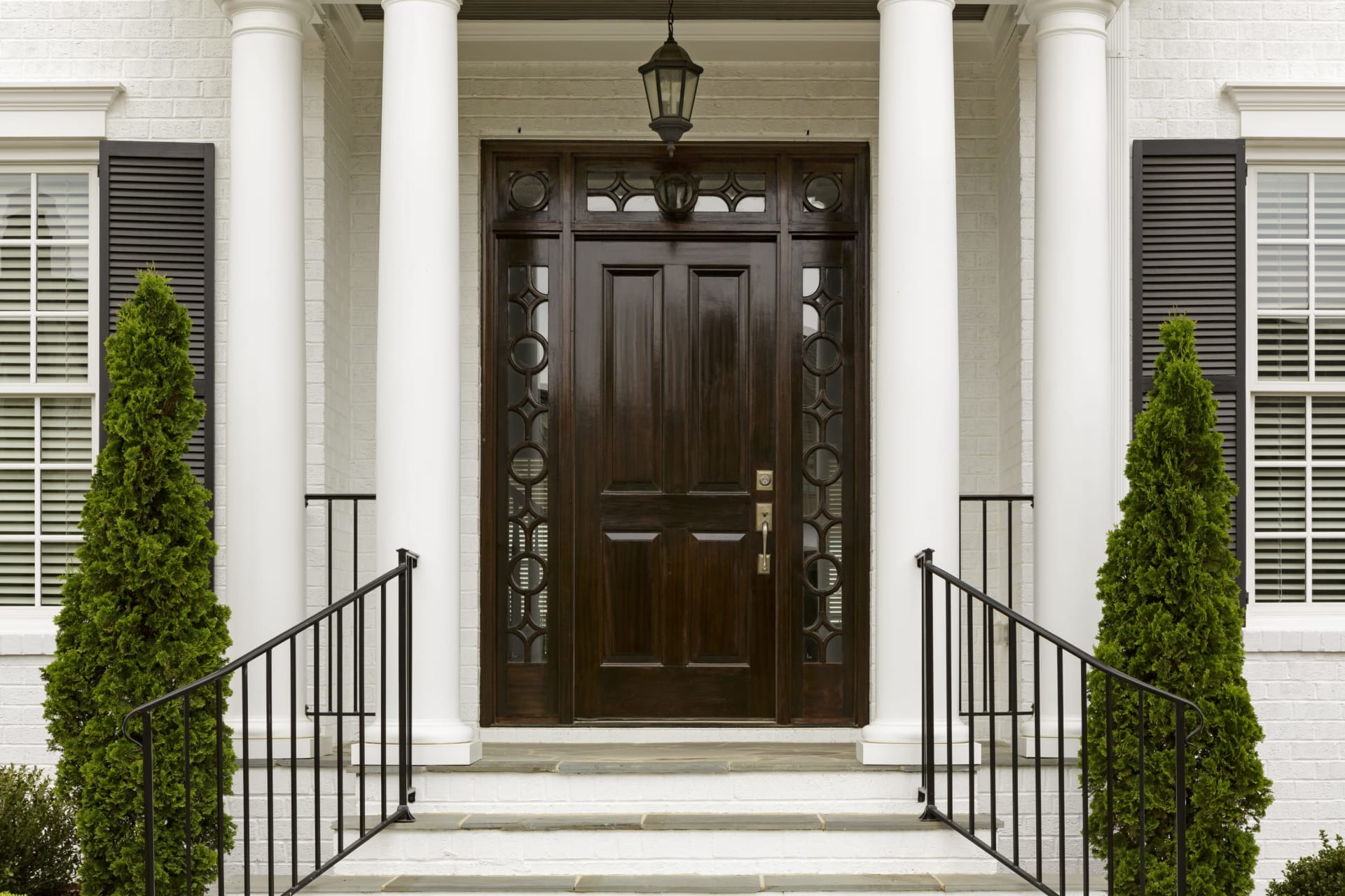 A modern white house with a close-up of the front wooden door, flanked by wrought iron railings, and a concrete pathway.