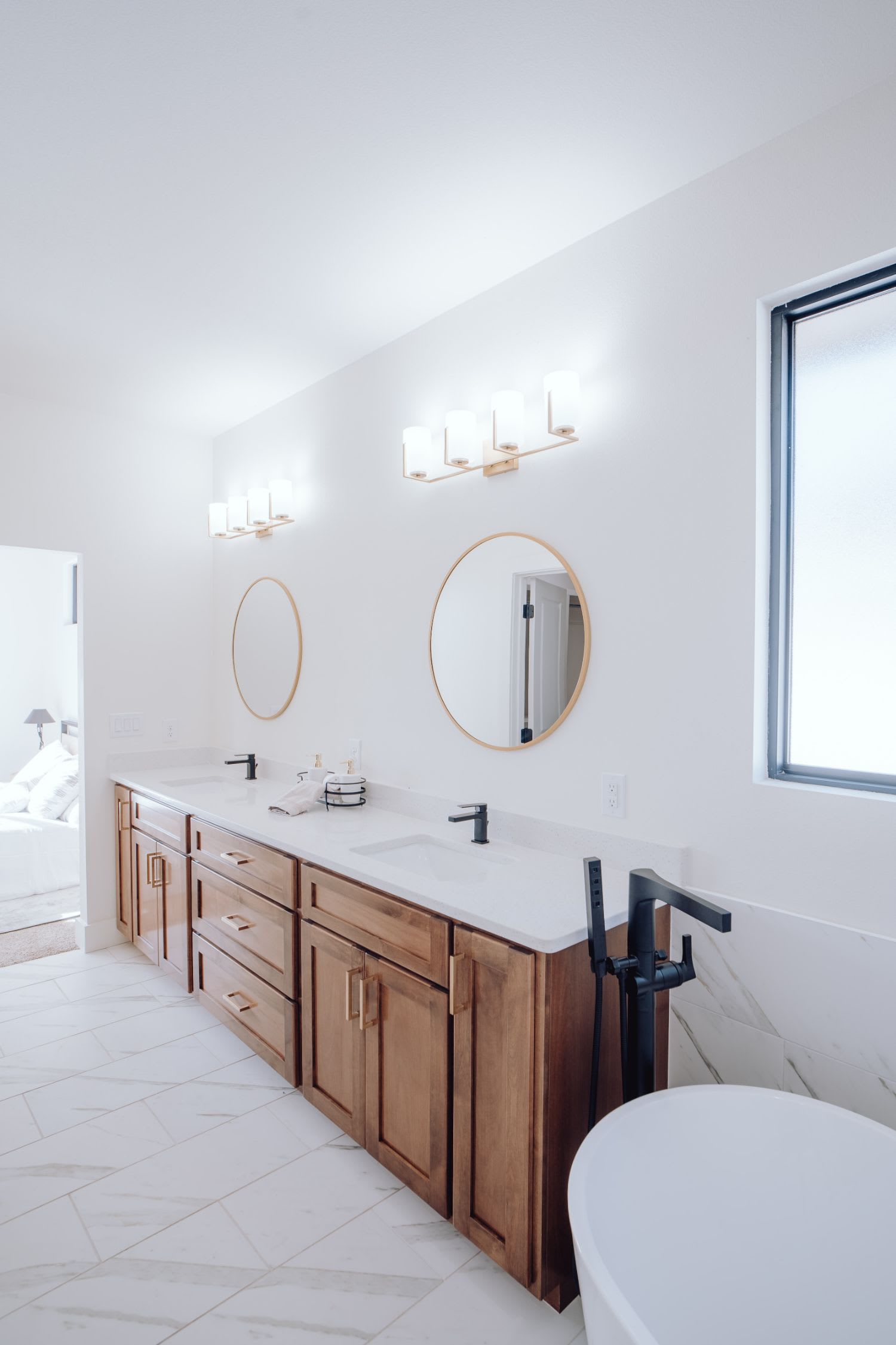 A bright bathroom with two sinks, a bathtub, a cabinet with drawers, mirrors, a window, and a bedroom view.