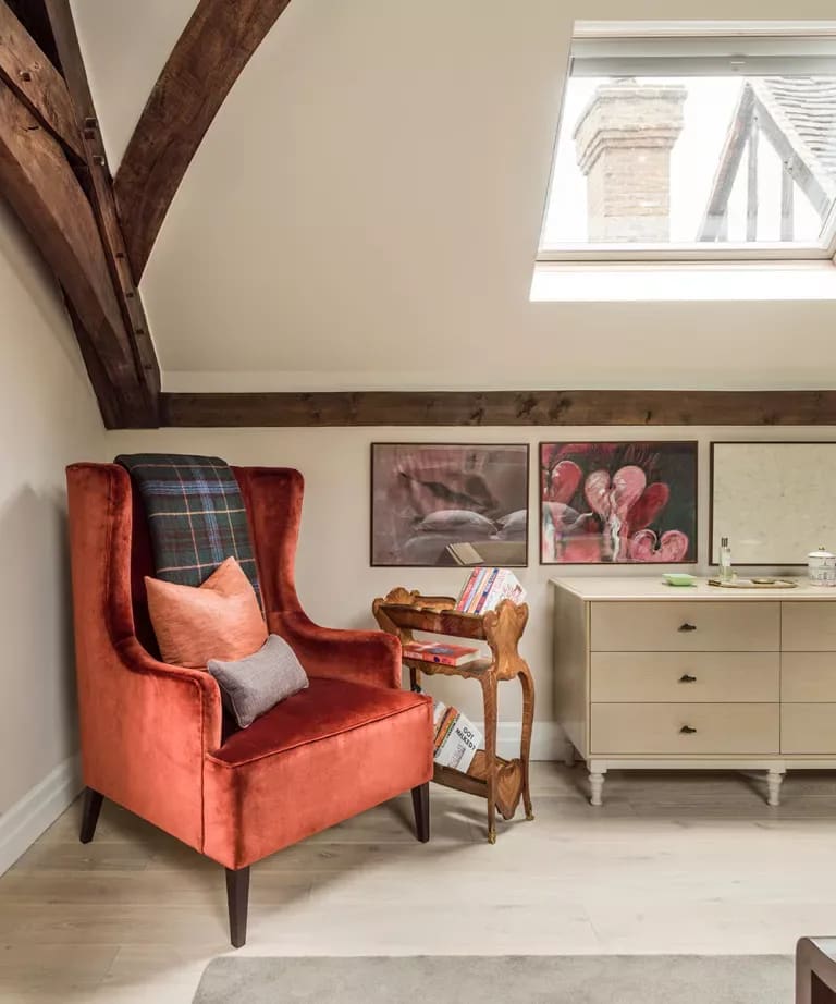 Rust-orange velvet wingback chair beside a cream chest of drawers beneath exposed beams and a skylight.
