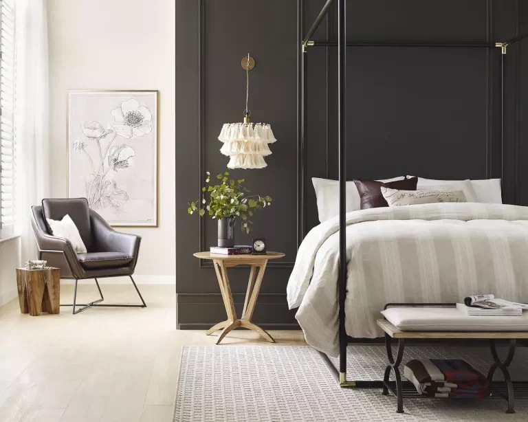 Staged bedroom with a black four-poster bed, charcoal wall, gray leather chair, and floral artwork.