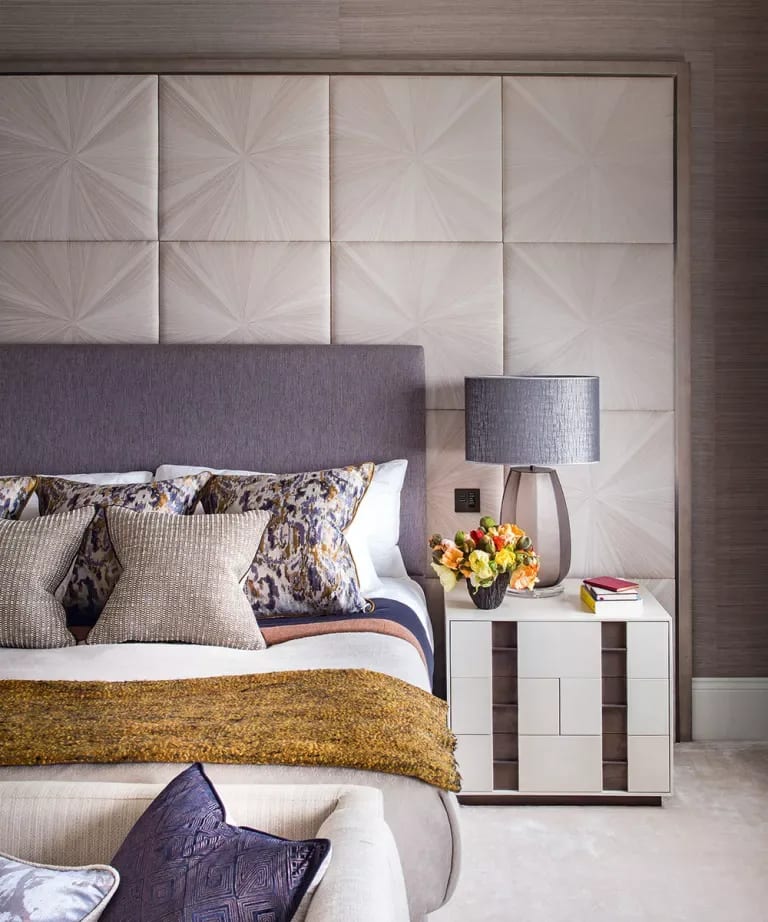 A styled bedroom with a geometric upholstered headboard, layered pillows, mustard throw, and bedside lamp.