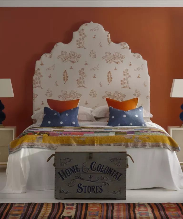 Bedroom with a patterned scalloped headboard, rust-colored walls, colorful bedding, and a vintage trunk.