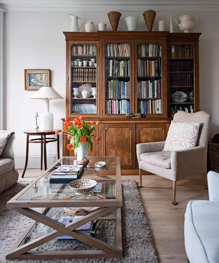 Living room with a tall wooden bookcase, pale armchairs, glass coffee table, and orange tulips.