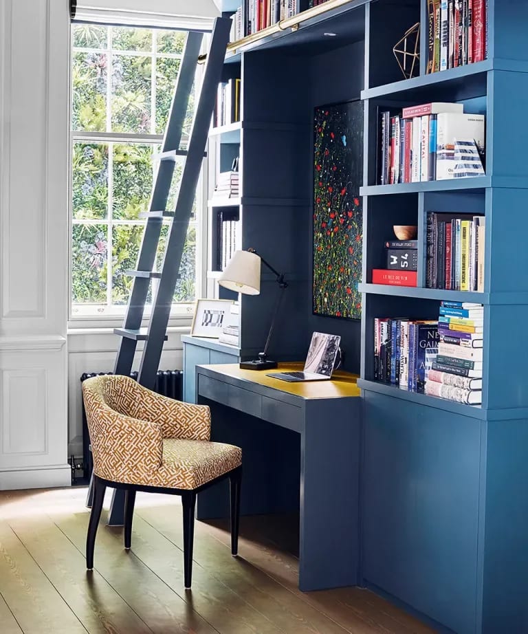 Home office with blue built-in bookshelves, a library ladder, and a patterned armchair beside a tall window.