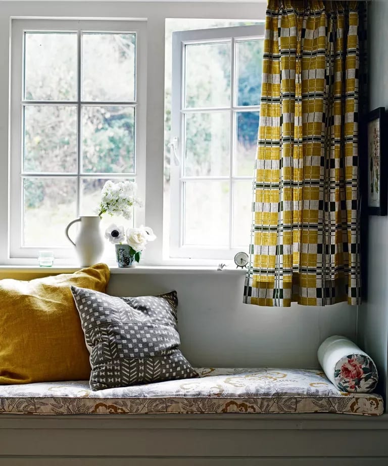 Window seat with mustard and patterned cushions beneath bright windows and yellow checked curtains.