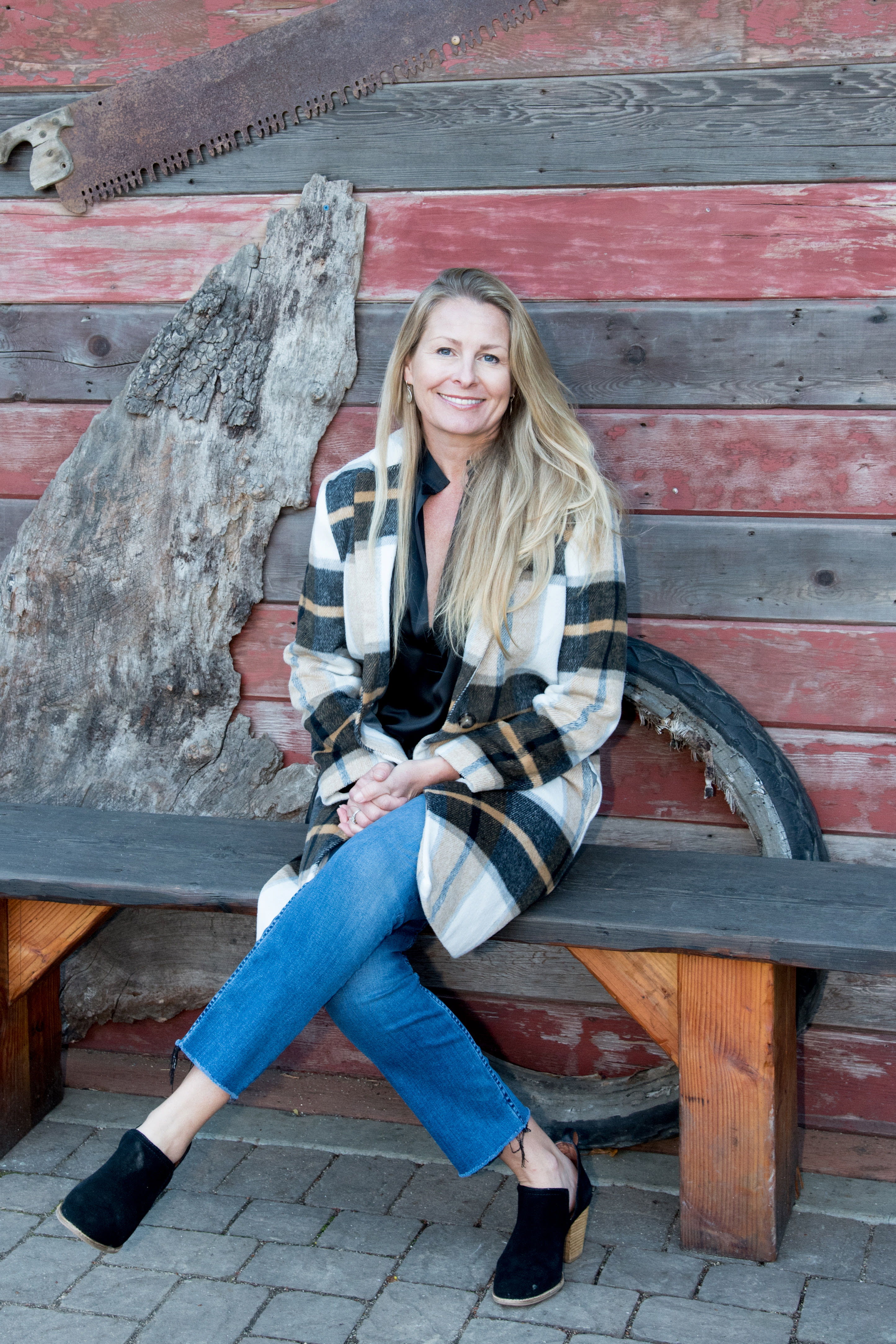 Smiling blonde woman seated on a bench in a plaid coat and blue jeans.