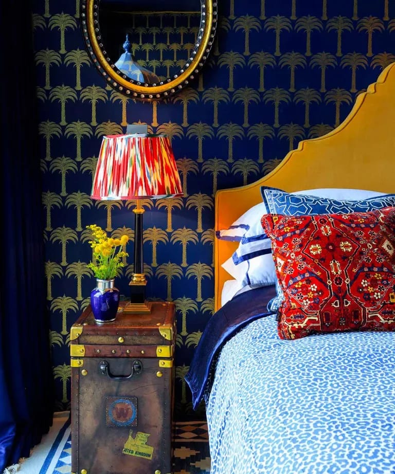 Bedroom with gold palm-patterned navy wallpaper, a mustard headboard, colorful pillows, and a vintage trunk nightstand.